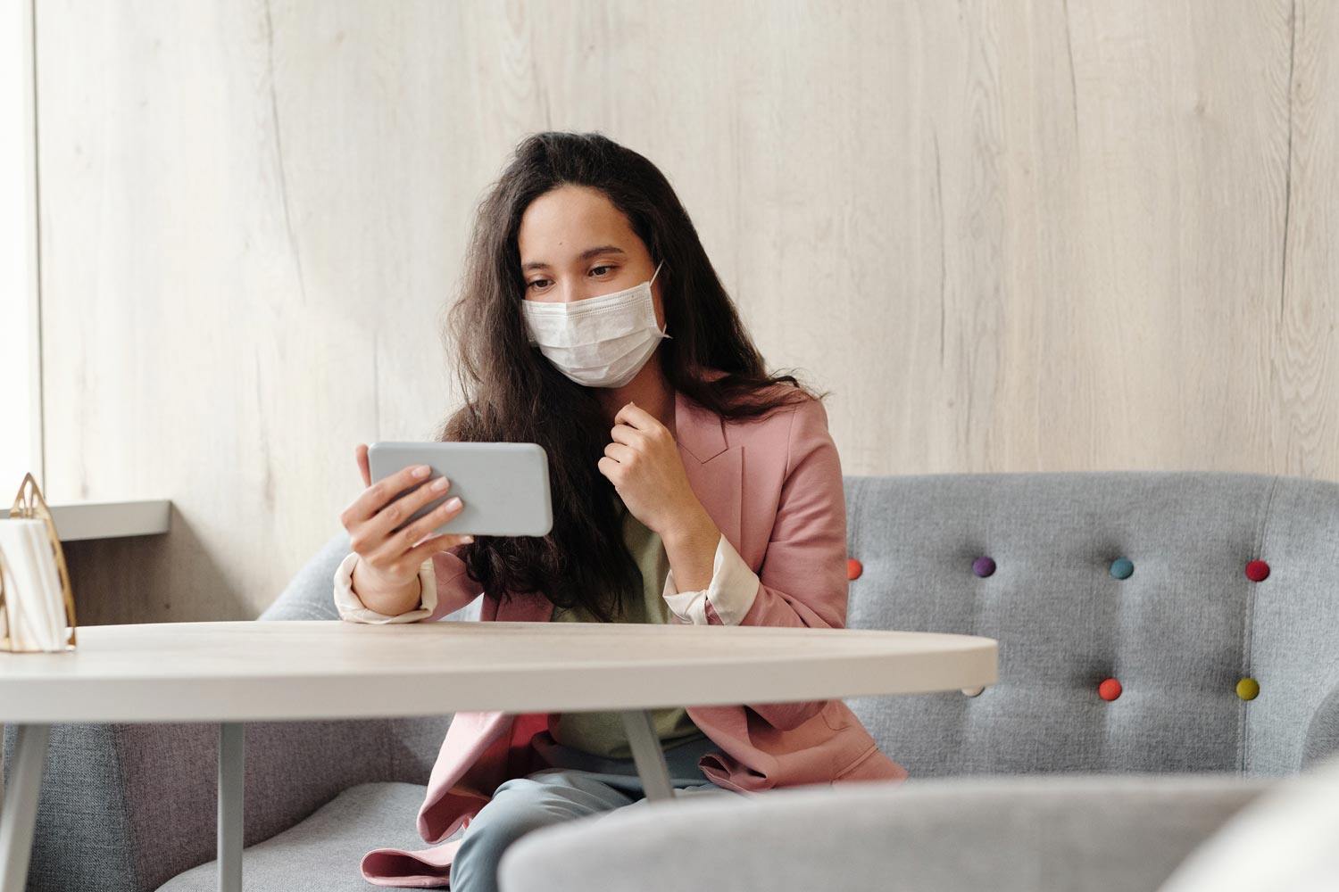 woman sitting in workplace with facemask, while on video chat on her cell phone